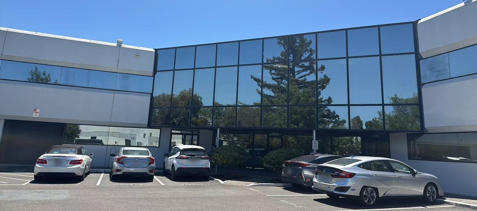 The Homebot One Inc office in Fremont, California: a two-story building with a wall of blue glass windows reflecting a tall pine tree.