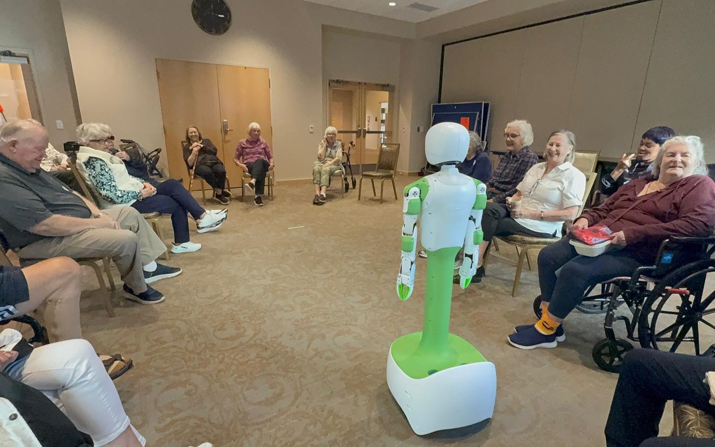 KOKO stands in the middle of a circle of smiling older adults, some in wheelchairs, in a senior living community’s activity room.
