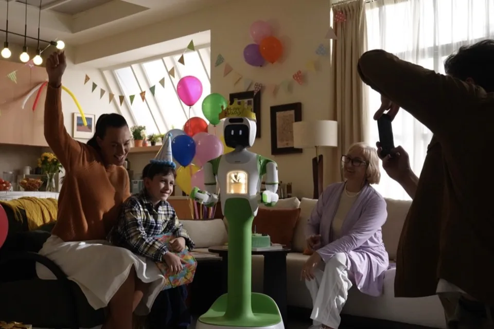 A family celebrates at home with balloons while KOKO stands beside them in the living room.