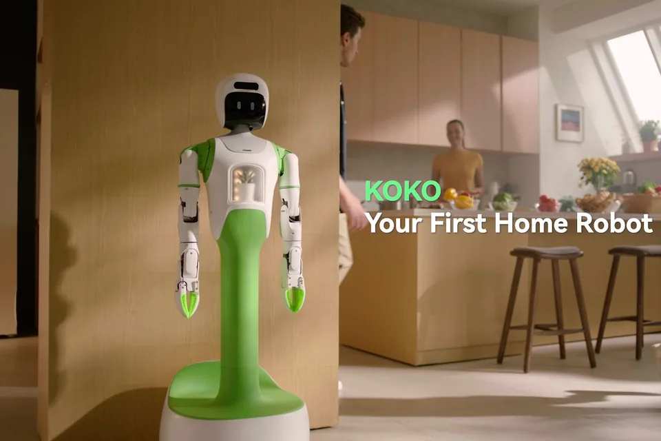 KOKO, a white and green home robot with two arms, stands in a sunlit kitchen while a family moves around it.
