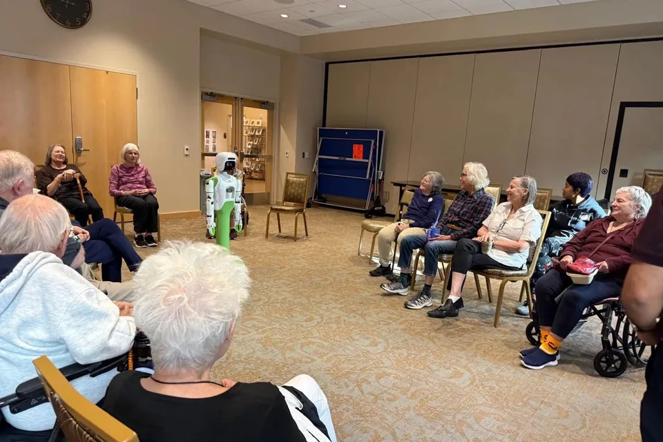 Older adults seated in a circle in a senior living community’s activity room, smiling as KOKO stands across the room.