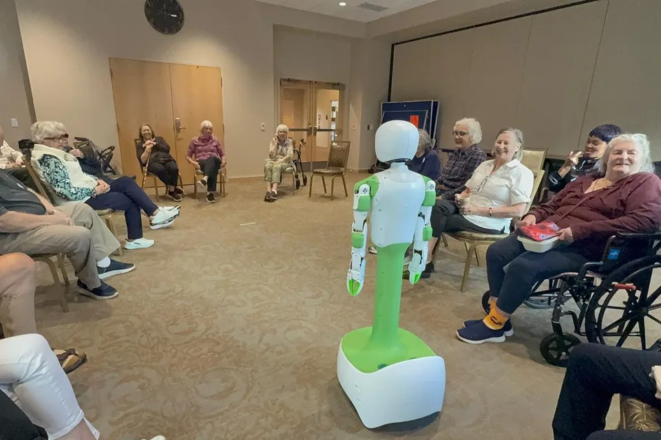 KOKO stands in the middle of a circle of smiling older adults, some in wheelchairs, in a senior living community’s activity room.
